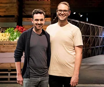 Two smiling men standing indoors, with fresh produce visible in the background.