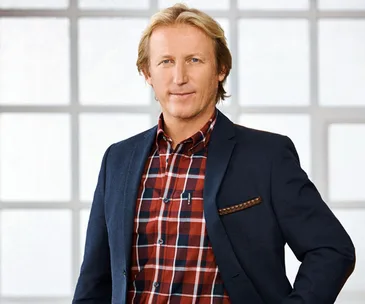 A person in a blue blazer and red plaid shirt stands in front of a large window.