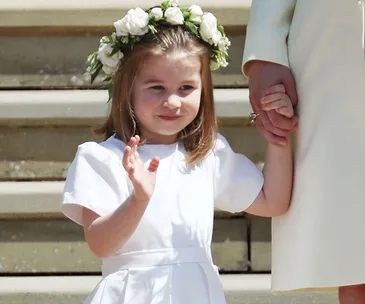 Young girl in white dress and flower crown holds an adult's hand, standing on steps and waving gently.
