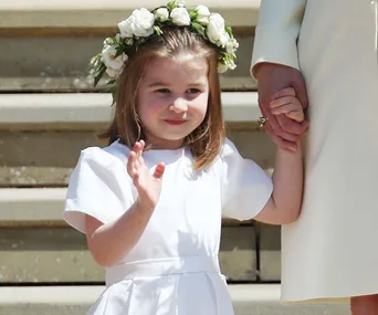 Young girl in white dress and flower crown holds an adult's hand, standing on steps and waving gently.