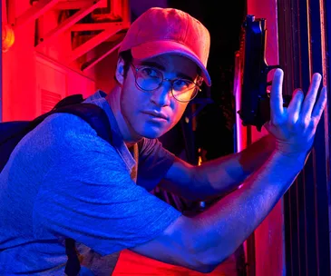 Darren Criss as Andrew Cunanan in a tense scene from "The Assassination of Gianni Versace," holding a gun and wearing glasses.