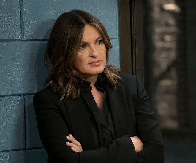 Detective in dark suit with arms crossed, standing against a blue wall, appears contemplative.