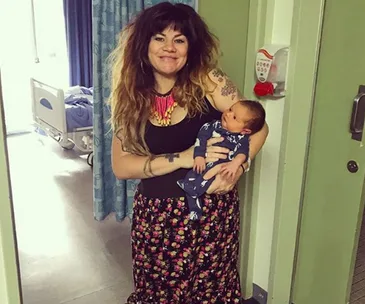 Woman smiling, holding a newborn in a hospital hallway, wearing a floral skirt and a colorful necklace.