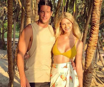 Two people standing on a sunlit beach, embracing, surrounded by trees.