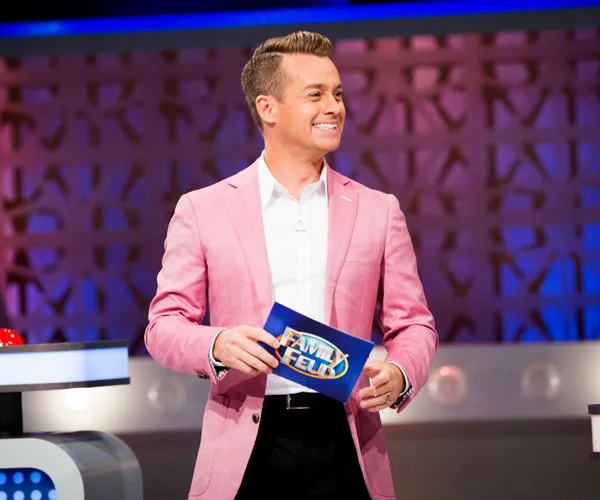 TV host in a pink blazer on the "Family Feud" set, holding a blue card.