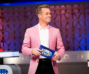 TV host in a pink blazer on the "Family Feud" set, holding a blue card.