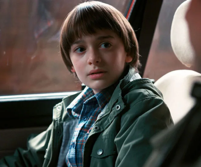 A young boy with brown hair and a green jacket sits in a car, looking concerned.