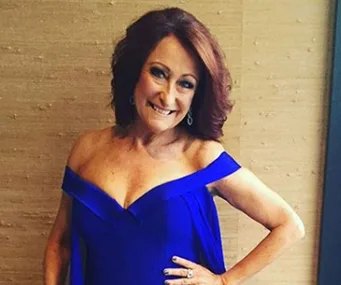 Lynne McGranger in an off-shoulder blue dress, smiling with hands on hips, against a beige background.