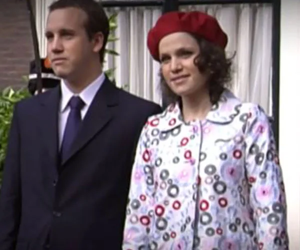 A man in a suit stands next to a woman wearing a floral coat and red beret, outdoors.