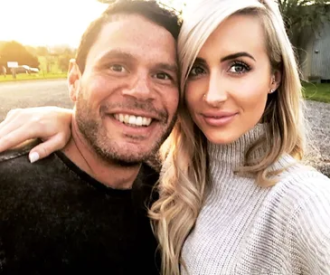 A smiling couple takes a selfie outdoors during golden hour, with the woman wearing a light turtleneck.