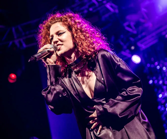 Singer with curly hair performing energetically on stage in purple lighting.