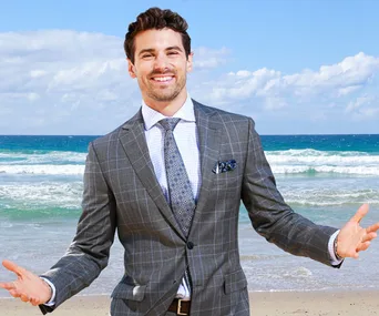 Man in a gray suit smiling with open arms on a beach, blue ocean waves in the background.