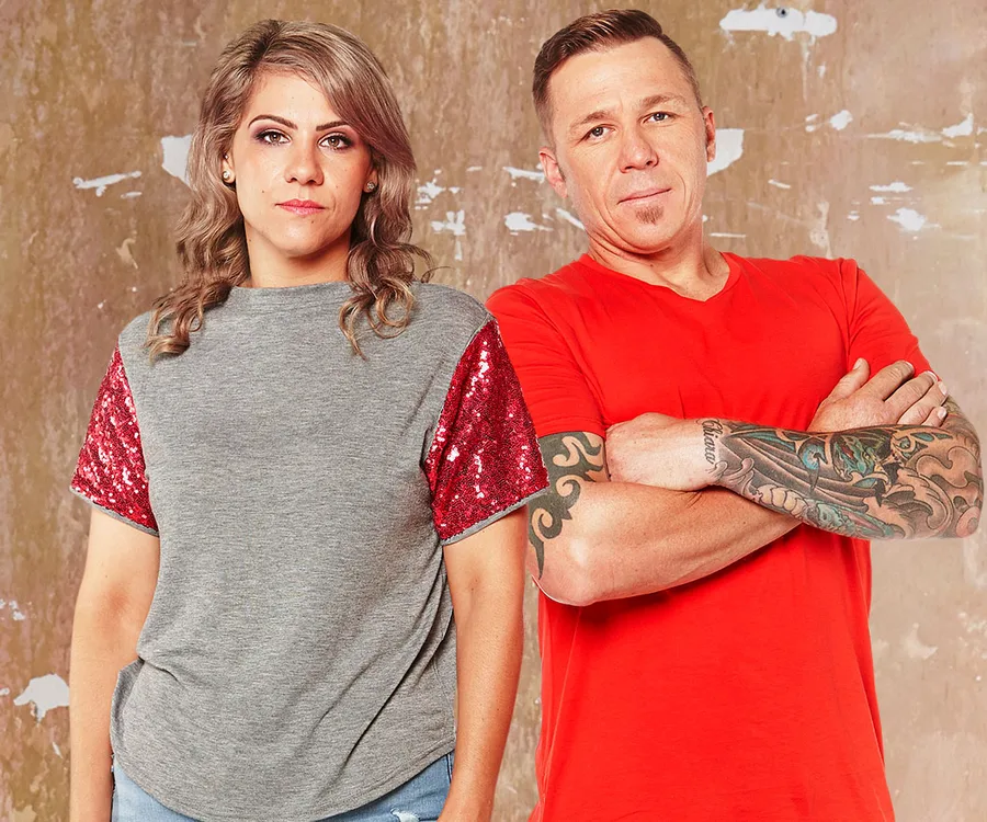 Two individuals stand side by side against a textured wall; one wears a gray shirt with red sleeves, and the other a red shirt.