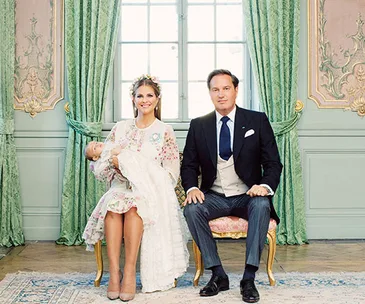 A woman holds a baby next to a man in a formal setting with ornate green curtains in the background.