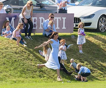 Woman in a light blue dress running on grass near children at a polo event with cars and onlookers in the background.