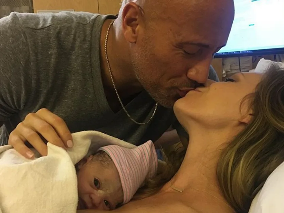 Man kissing woman on hospital bed holding newborn baby peeking from a blanket.