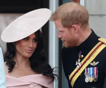 Prince Harry and Meghan Markle on a palace balcony, engaged in conversation, with Meghan wearing a pink hat.