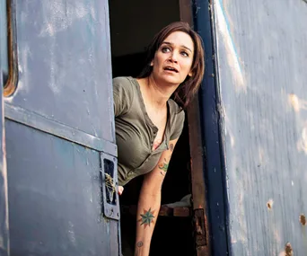 A woman with tattoos leans out of a blue metal door, looking ahead with a concerned expression.