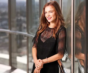 A woman in a sheer black dress smiles while leaning on a glass railing, with a cityscape view behind her.