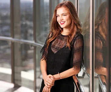 A woman in a sheer black dress smiles while leaning on a glass railing, with a cityscape view behind her.