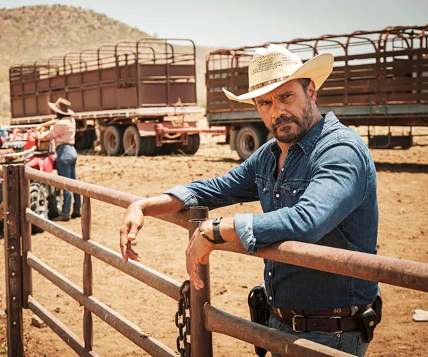 Male cowboy in denim and hat leans on a gate with cattle trucks in background, in a rural setting from "Mystery Road."