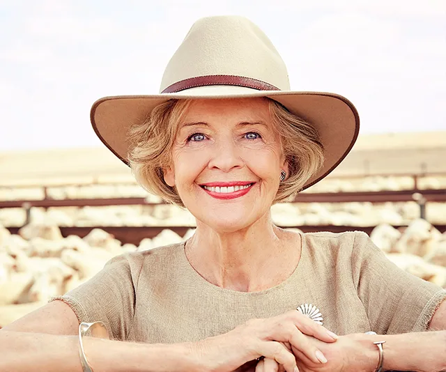 Elderly woman smiling outdoors, wearing a beige hat and top, with sheep in the background.
