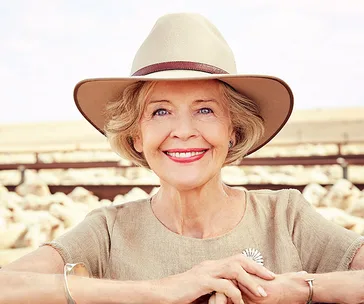 Elderly woman smiling outdoors, wearing a beige hat and top, with sheep in the background.