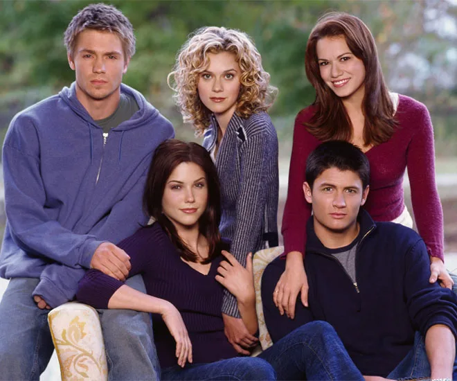 Group photo of five young adults from "One Tree Hill," smiling outdoors in casual clothing.