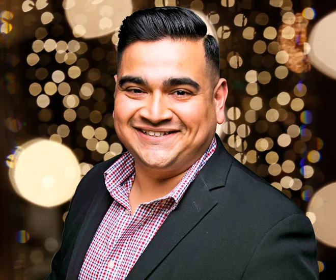 A smiling man in a suit against a festive, blurred lights background.