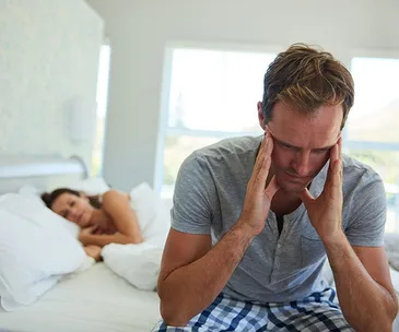 Man sitting on bed looking distressed, woman lying in bed in the background, appearing concerned.