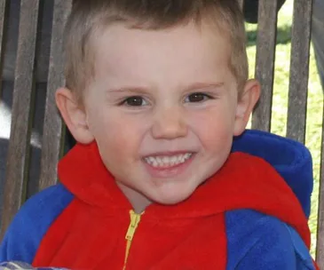 A young child wearing a red and blue hooded outfit smiles while sitting outside on a sunny day.