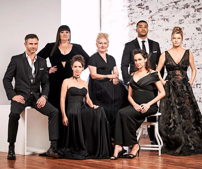 Cast members of Wentworth Season 6 pose elegantly in formal black attire against a white brick backdrop.