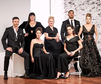 Cast members of Wentworth Season 6 pose elegantly in formal black attire against a white brick backdrop.