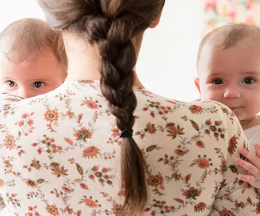 A woman with a braid holding twin babies, wearing a floral shirt, facing away while the babies look forward.