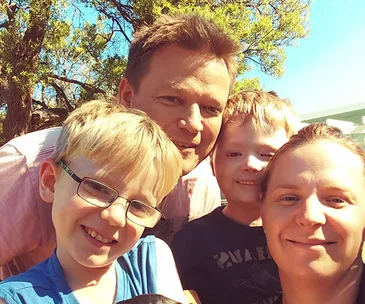 A smiling family outdoors under a tree with clear blue sky.