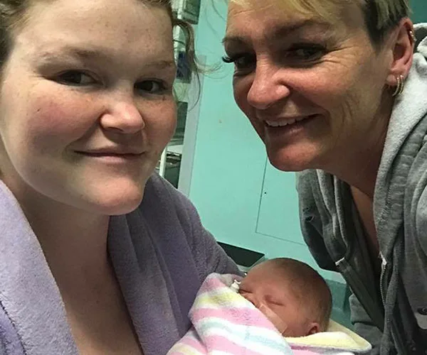 Mother and another woman smiling at the camera while holding a newborn wrapped in a striped blanket.
