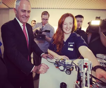 A woman with red hair demonstrating a robot to a man in a suit, surrounded by photographers.