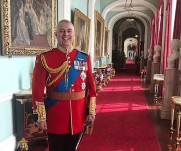 Man in red military uniform stands in an ornate hallway with red carpet and portraits on the walls.