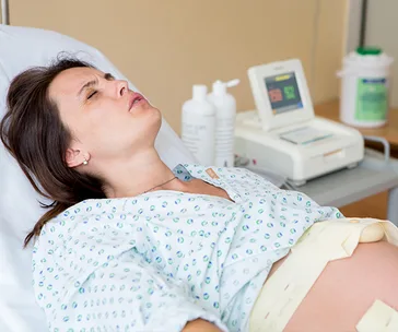 Pregnant woman in hospital gown on bed with a fetal monitor and medical equipment nearby, appearing to be in labor.
