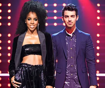 Two people standing on stage with colorful lights in the background, one wearing a black sequined outfit, the other in a dark suit.