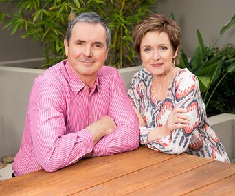 A man in a pink striped shirt and a woman in a patterned blouse sit together outdoors at a wooden table.