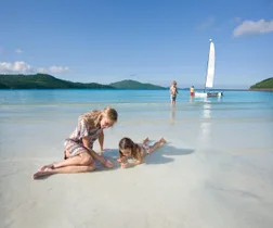 Mother and child playing on sandy beach, with children and a sailboat in shallow blue water, green hills in background.