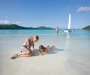 Mother and child playing on sandy beach, with children and a sailboat in shallow blue water, green hills in background.