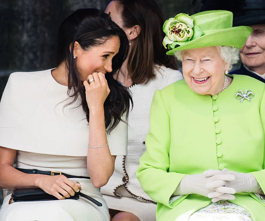 Two women seated, one in a white dress and the other in a bright green outfit and hat, smiling and engaging with each other.