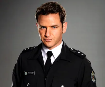 Actor in uniform from TV series "Wentworth", wearing a black officer's jacket and name tag against a gray background.