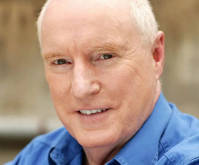 Portrait of a man in a blue shirt, smiling softly, with a blurred neutral background.