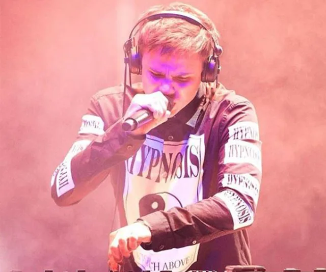 Young man wearing headphones performing on stage with a microphone, pinkish lighting ambiance.