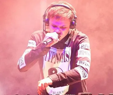 Young man wearing headphones performing on stage with a microphone, pinkish lighting ambiance.
