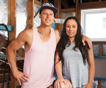 Couple posing in a partially renovated room, with wooden beams and insulation visible in the background.