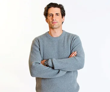 Man in a gray sweater with arms crossed against a plain background.